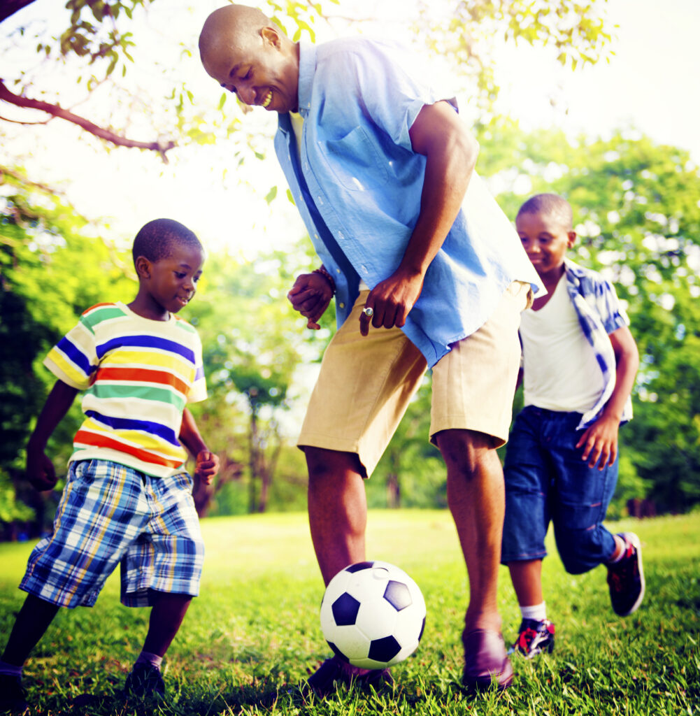 An adult and two children play soccer on grass in a park on a sunny day, all smiling and wearing casual summer clothes.