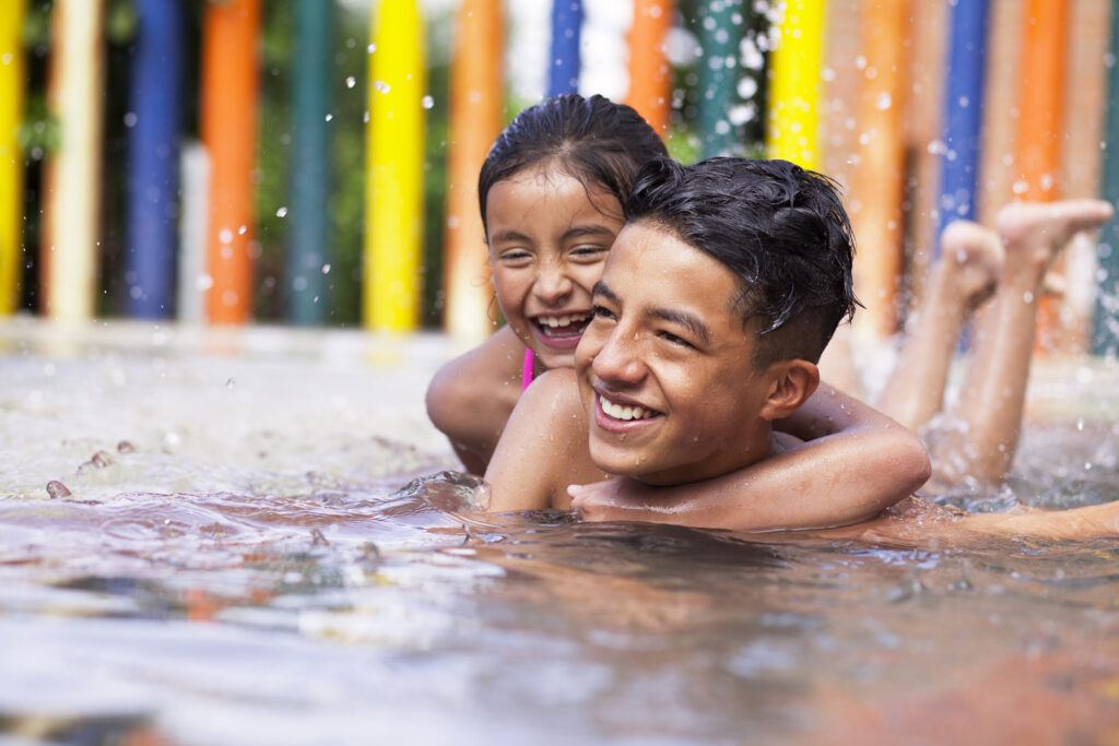 Two smiling children play and swim in shallow water, with colorful vertical poles in the background and water splashing around them.