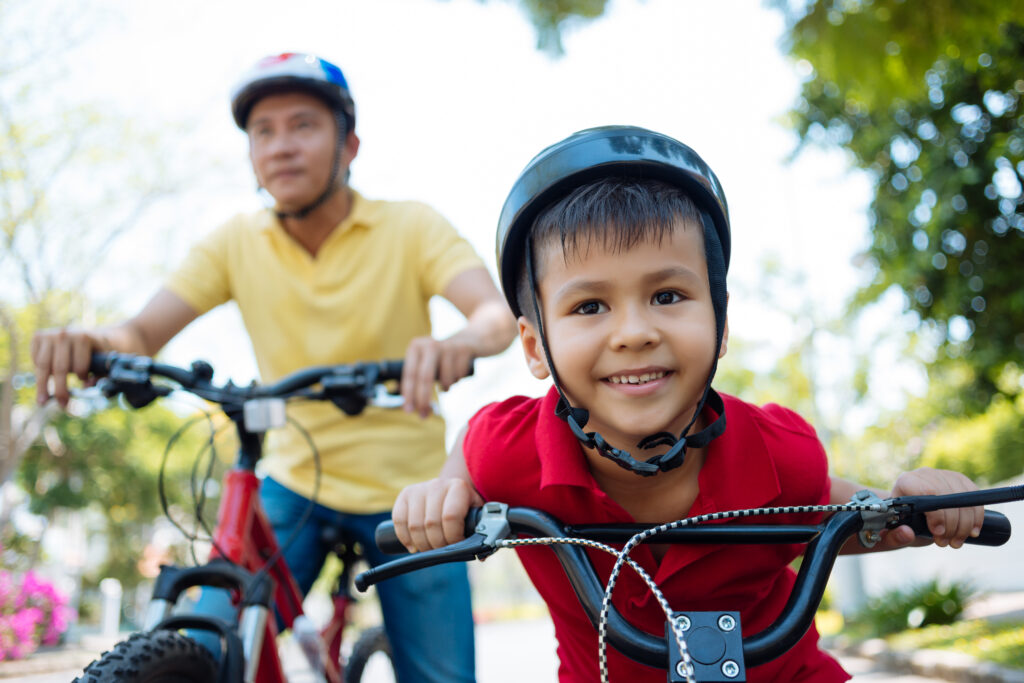 A young boy in a helmet riding a bike in the foreground, with an adult in a yellow shirt biking behind him outdoors.